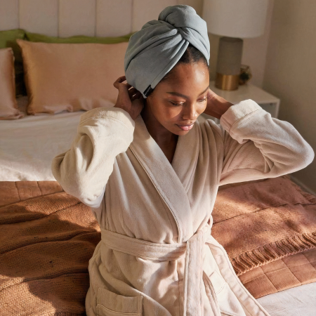 Woman sitting on her bed wearing a beige robe and head wrap, relaxing after skincare, surrounded by silk bedding—showcasing the benefits of Sillo silk pillowcases for radiant, hydrated skin.