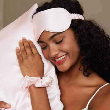 Woman resting on a blush pink Sillo silk pillowcase while wearing a matching silk eye mask and scrunchie, showcasing smooth, frizz-free hair and the natural sheen of 22-Momme mulberry silk that supports scalp and hair health.