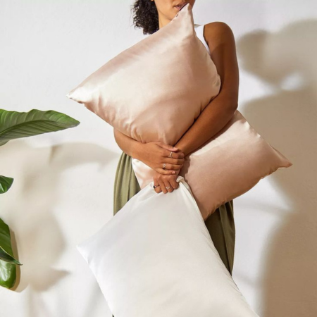 Woman holding champagne and ivory Sillo silk pillowcases, illustrating the softness, natural sheen, and luxurious care of 22-Momme mulberry silk designed to stay smooth and durable with proper washing and drying.