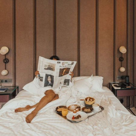 Traveler relaxing in bed with a breakfast tray, reading a newspaper beside a Sillo silk pillowcase — a silver-infused, hypoallergenic silk pillowcase that prevents breakouts and frizz while maintaining skin hydration during travel.