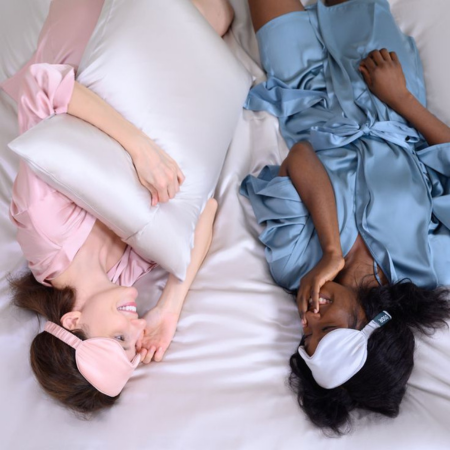 How silver ions keep your pillowcase bacteria-free and promote clearer, healthier skin. Image shows two women relaxing on a bed wearing silk robes and eye masks, chatting and smiling with Sillo silk pillowcases beneath them