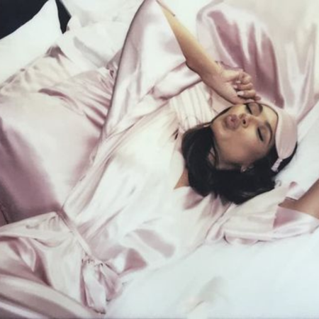 Woman wearing a pink silk sleep mask and robe relaxing in bed with Sillo’s silver-infused silk pillowcase — showing how silk’s smooth, antibacterial surface helps reduce acne, irritation, and dryness for clearer skin overnight.