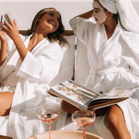 Two women in white robes enjoying a self-care moment with silk pillowcases and skincare masks — promoting youthful, hydrated skin and wrinkle prevention.