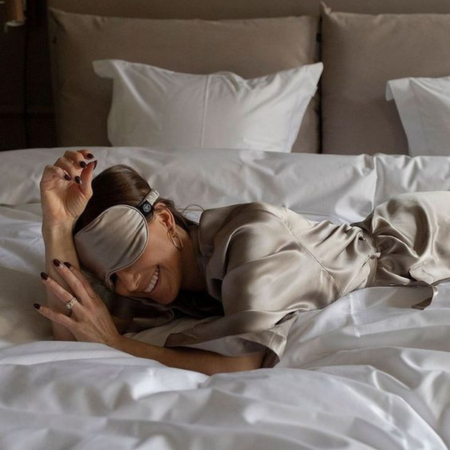Image shows a woman relaxing on a bed in a silk eye mask, resting on a soft ivory Sillo silk pillowcase that protects hair from friction and dryness.