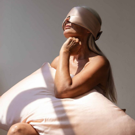 Mature woman resting with a champagne Sillo silk pillowcase and matching sleep mask — illustrating how silk supports smoother skin and healthier hair overnight.