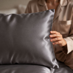 A close-up of a person in matching beige pajamas holding charcoal grey Sillo silk 22 momme mulberry silk pillowcase with envelope closure  in a softly lit bedroom setting.
