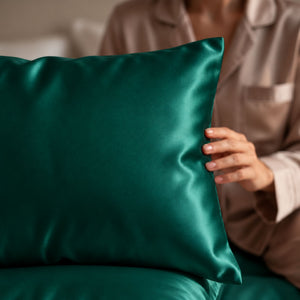 A close-up of a person in matching beige pajamas holding emerald green Sillo silk 22 momme mulberry silk pillowcase with envelope closure  in a softly lit bedroom setting.