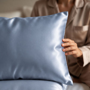 A close-up of a person in matching beige pajamas holding Haze Blue Sillo silk 22 momme mulberry silk pillowcase with envelope closure  in a softly lit bedroom setting.