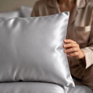 A close-up of a person in matching beige pajamas holding silver Sillo silk 22 momme mulberry silk pillowcase with envelope closure  in a softly lit bedroom setting.