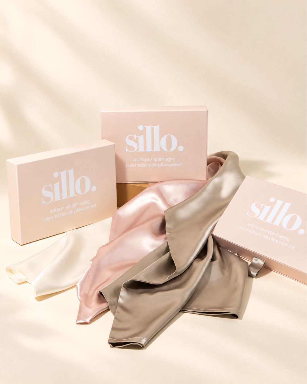 Three Sillo silk pillowcase gift sets in ivory, blush pink, and light champagne made with 22 momme mulberry silk woven with silver ions, draped over their boxes, a silk pillowcase gift for skin and hair. 