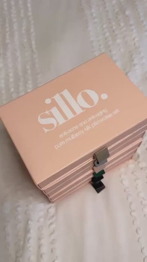 Unboxing video of the Sillo silk pillowcase gift set: a blush pink Sillo gift drawer box opens to reveal two 22 Momme Grade 6A mulberry silk pillowcases woven with silver ions and a Sillo Care Washbag, with a stack of gift boxes showing shade tabs on white bedding. 